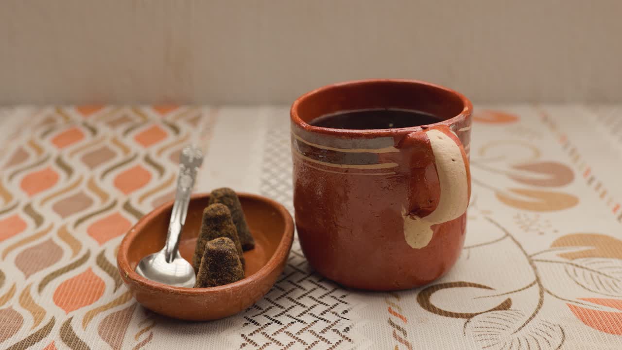 Clay mug with black coffee and a small dish holding brown sugar (piloncillo) and spoon on a patterned cloth