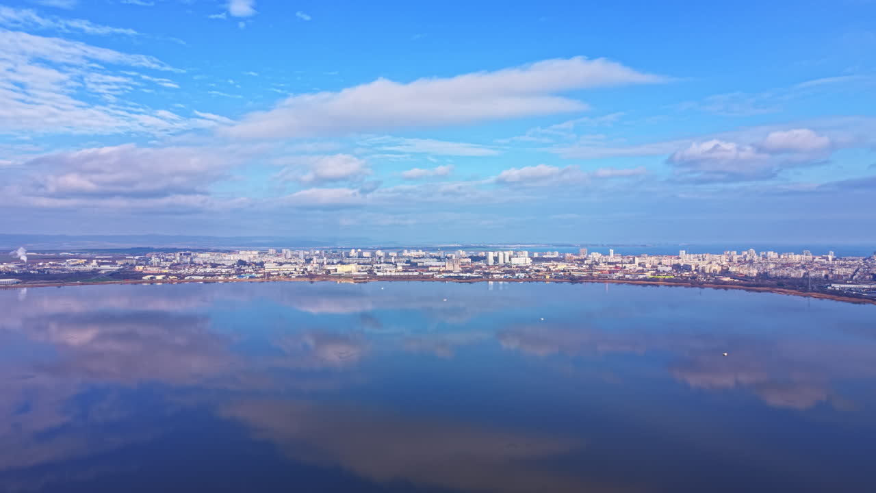 晴れた日、大湖のほとりの街。空と水面に建物や雲が映り込み、地平線の上下には様々な青のグラデーションが広がっている。