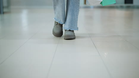 front leg view of person wearing frayed jeans and chunky boots walking in mall carrying shopping bags, soft shadow cast on polished tile floor, urban structure visible in blurred background