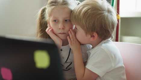 Brother-tells-sister-secret-sitting-at-table-with-laptop