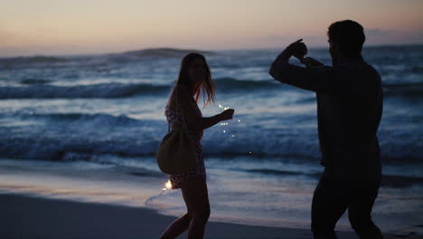 Fun-couple,-sparkles-and-phone-on-sunset-beach