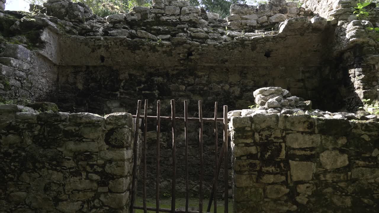 Stone ruins showcase weathered walls, wooden bars blocking an entrance, moss covers the stones, archaeological site in a forested area, sunlight highlights the textures.
