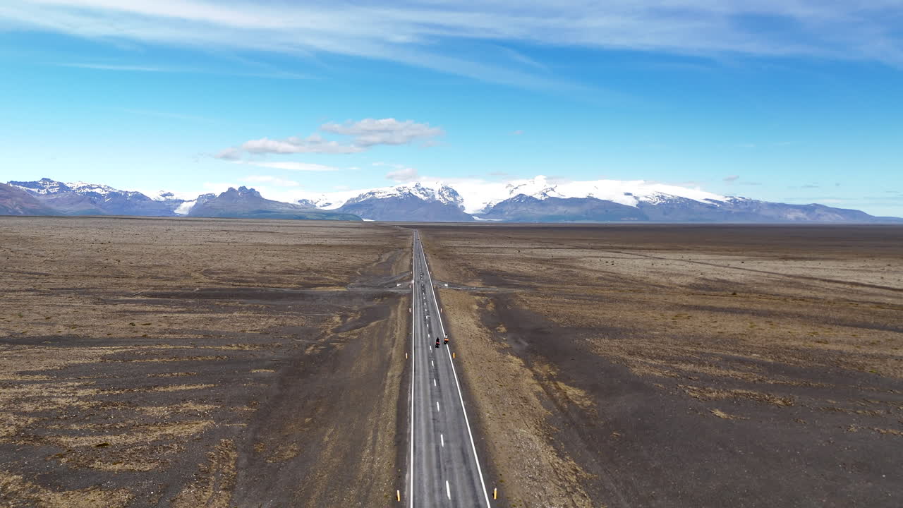 Vista aérea de drone de dois ciclistas pedalando em uma estrada remota perto da geleira Vatnajökull, na Islândia, atravessando uma vasta planície vulcânica com montanhas cobertas de neve sob um céu azul claro