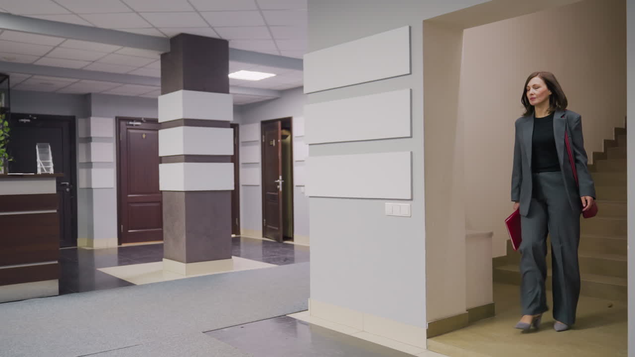Businesswoman in formal attire walking down office stairwell carrying folder, professional office interior, modern workplace, confident woman in business setting