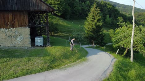 Professional-gardener-mowing-lawn-near-a-wooden-cottage-with-gasoline-brush-cutter,-parallax-shot