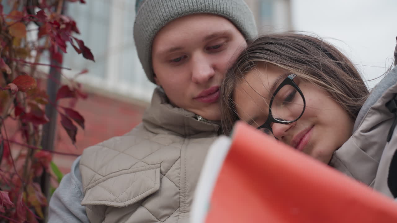 Lovely woman rests her head on husband chest as hair flutters gently in wind, both warmly dressed and reading book together outdoors, with blurred background featuring autumn leaves