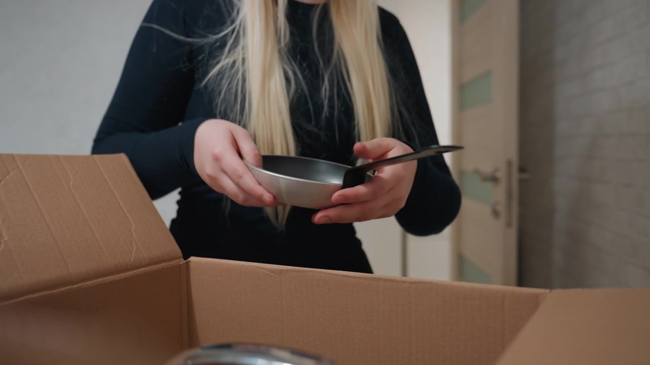 White woman unpacking cardboard box plates, long blonde hair over black sweater, hands carefully lifting ceramic dish and cork trivet, placing items on kitchen shelf near apartment door, soft natural
