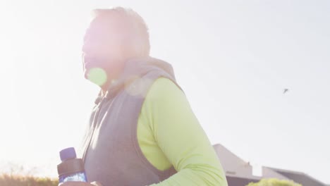 Video-Eines-älteren-Gemischtrassigen-Mannes-In-Sportkleidung-Und-Kopfhörern,-Der-Auf-Einer-Sonnigen-Straße-Läuft