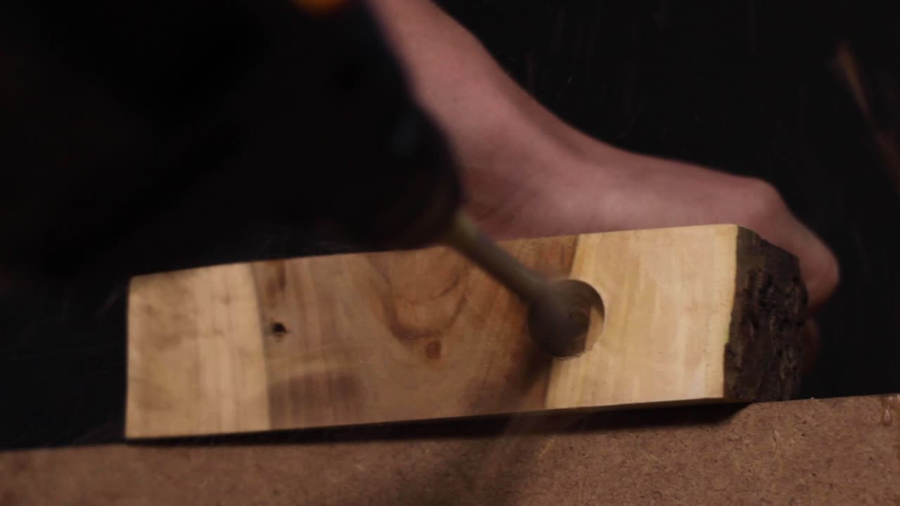 Close up of a carpenter using a special drill to create a large hole in a thick slab of processed, smooth wood, holding it steady with his hand