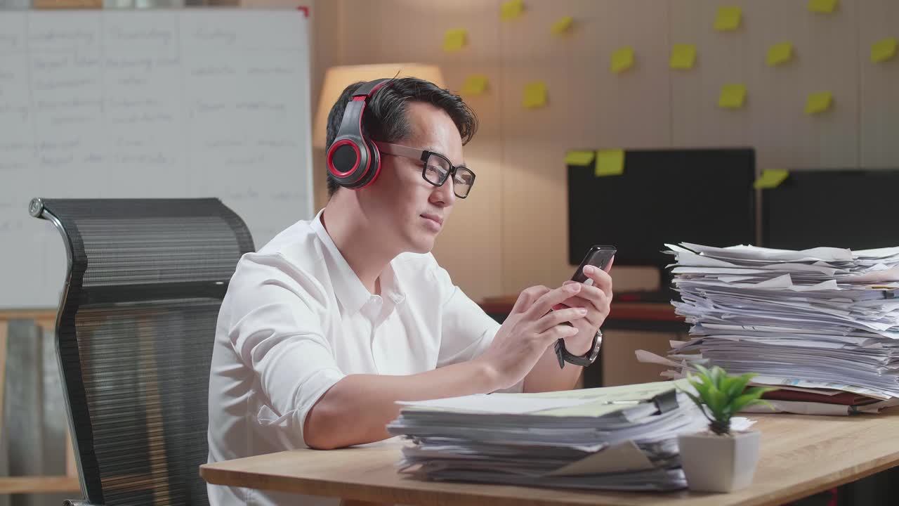 Asian Man With Headphones Listening To Music On Smartphone After Working With Documents At The Office