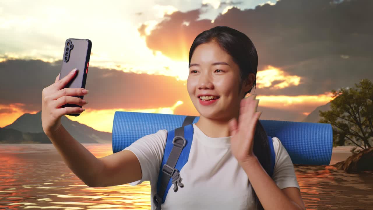 Close Up Of Asian Female Hiker With Mountaineering Backpack Waving Her Hand And Pointing Back While Having A Video Call On Smartphone At A Lake