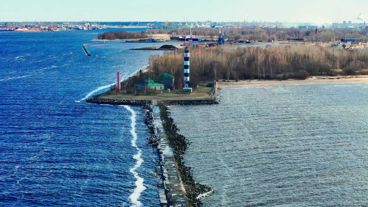 A tall black and white lighthouse stands at the end of a narrow stone jetty splitting vivid blue Baltic Sea from muted estuary waters, bordered by forest and distant industry.