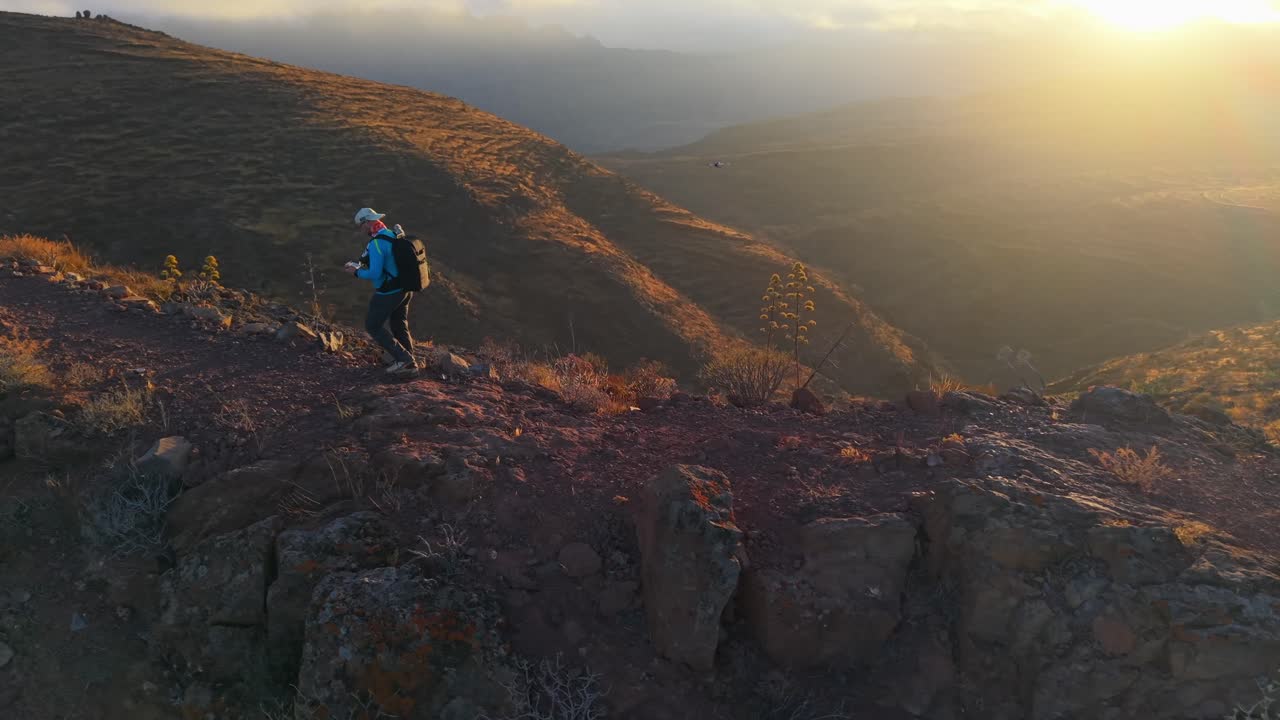 Man walking along a mountain trail and a drone tracking him at sunset