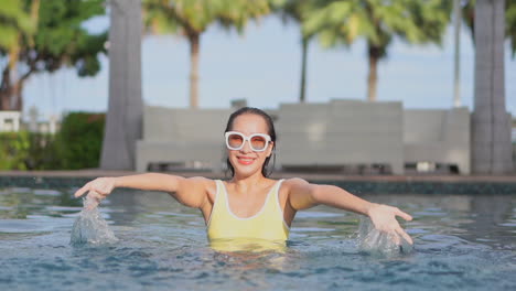 Fröhliche,-Attraktive-Frau-In-Leuchtend-Gelbem-Badeanzug-Und-Großer-Weißer-Sonnenbrille,-Die-Im-Pool-Planscht-Und-Lächelt,-Während-Sie-Im-Urlaub-An-Einem-Exotischen-Luxusziel-Ist
