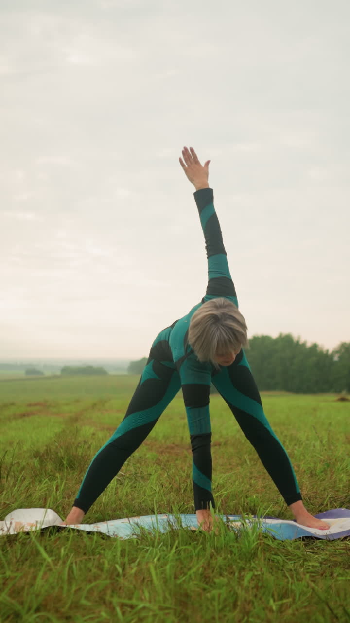 mujer en traje verde y negro en una alfombra de yoga practicando una postura triangular hacia adelante en un campo sereno de hierba bajo un cielo nublado con árboles en el fondo, brazos extendidos