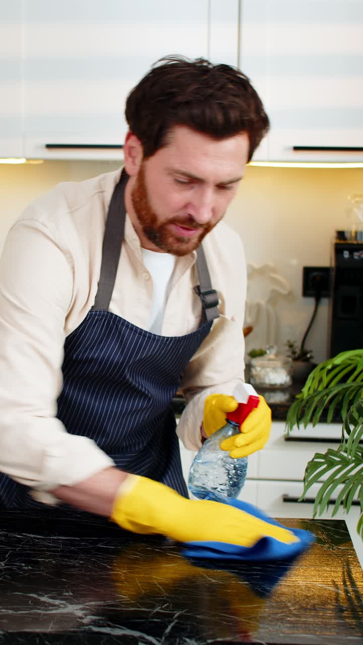 Uomo di mezza età che sorride mentre pulisce il tavolo della cucina con un panno per l'igiene e la freschezza quotidiana della casa.