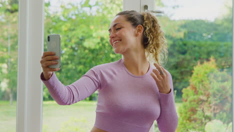 young woman sitting on yoga mat at home after exercise posing for selfie on mobile phone