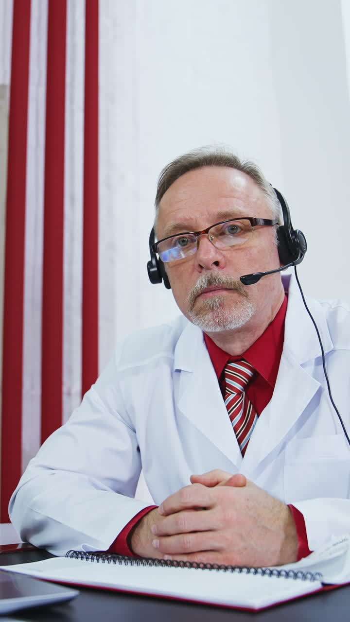 Older doctor listens to patient during video call. Senior male physician in medical uniform and headset having a remote consultation. Telemedicine concept. Vertical video