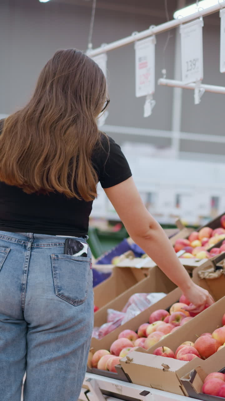 vista trasera de una mujer con el teléfono en el bolsillo trasero recogiendo una manzana roja de la exhibición, observándola y dejándola caer, etiquetas de precios y señales visibles en el fondo de un entorno de tienda brillante