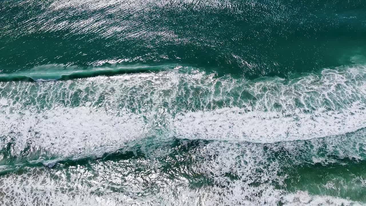 High angle drone shot captures turquoise ocean waves breaking over a sandy beach in Australia