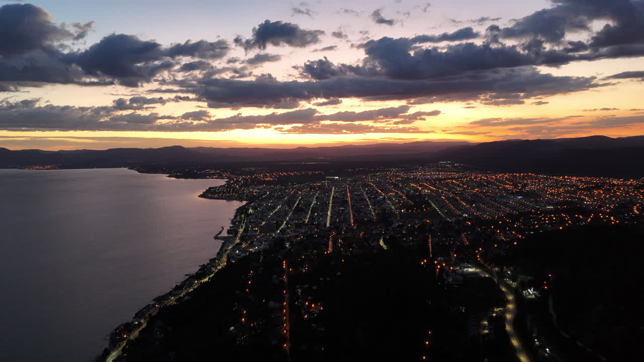 amanecer aéreo en bariloche, argentina, luces callejeras y coches iluminan la red de la ciudad mientras el sol brilla contra las nubes en el cielo