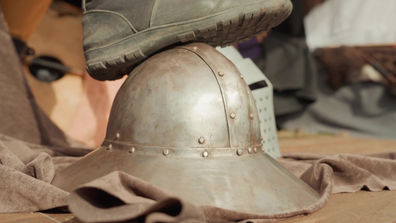Uno stivale preme un elmo di metallo usurato su un tessuto marrone piegato, inclinando la tesa e comprimendo il tessuto, mentre la cupola graffiata mostra rivetti, ammaccature, ruggine e una texture invecchiata sotto una calda luce direzionale.