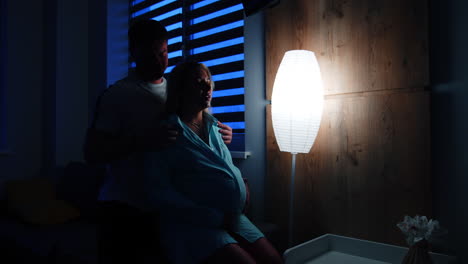 husband caressing his pregnant wife sitting on the bed. woman prepares for childbirth in hospital. dark room backdrop.
