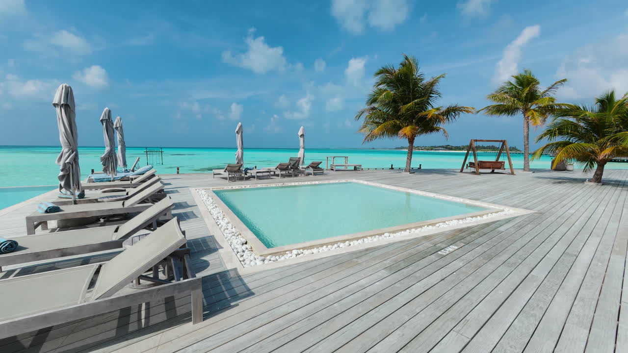 Push in shot over a luxury wooden deck at a tropical resort showing sun loungers a small pool palm trees and beautiful blue ocean water in the Maldives