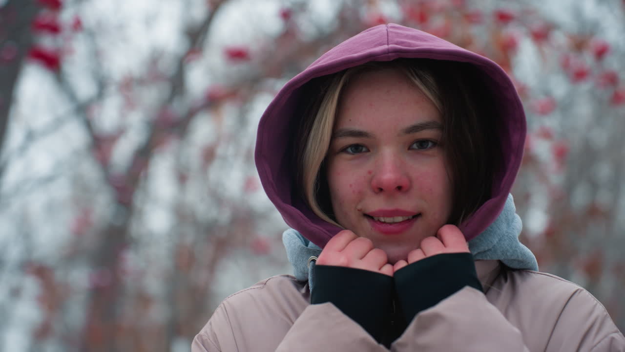 близкий взгляд на молодую женщину, слегка улыбающуюся, держащую воротник с капюшоном, глядя на камеру с тонкой улыбкой, зимняя куртка, наружная обстановка с размытыми деревьями на заднем плане