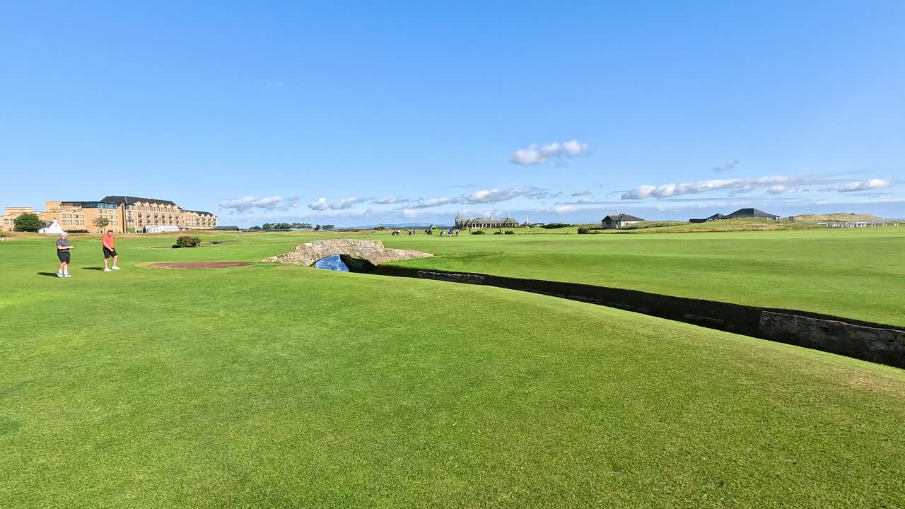 Two golfers walk across iconic stone bridge on sunny fairway, wide shot, steady camera