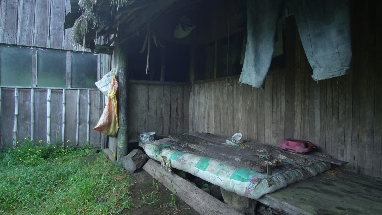 Static shot of a rustic wooden shelter in a rural environment, showing an old improvised bed, hanging clothes and weathered wooden walls