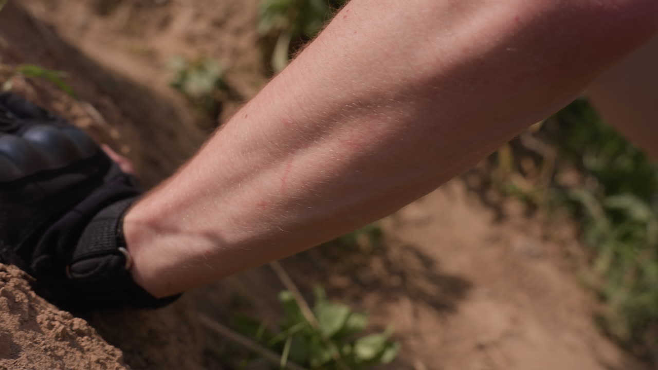 close up adventurer arms palms pressed firmly on steep grassy hill soil next to water filled hole while man performs push up in outdoor survival terrain showcasing strength focus endurance