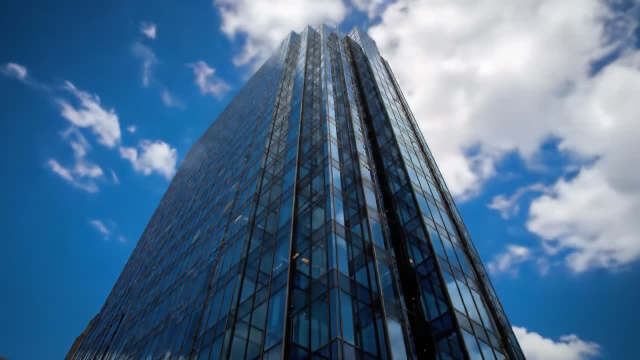 A tall modern building showcases its stunning glass exterior under a bright blue sky filled with fluffy clouds. The architecture highlights intricate design and urban beauty.