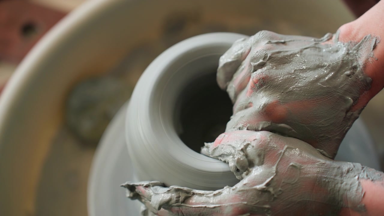 Closeup hands shaping clay on wheel, wet gray slip forming rounded bowl rim with gentle pressure, artisan fingers smoothing surface, studio light highlighting texture, slow rhythmic rotation conveys