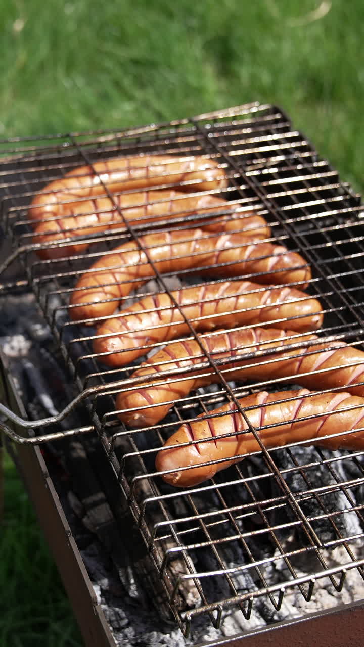 Roasting sausages on a grill outdoors. Delicious hot dog sausages grilling in smoke among green nature. Cooking meat sausages for a picnic. Vertical video