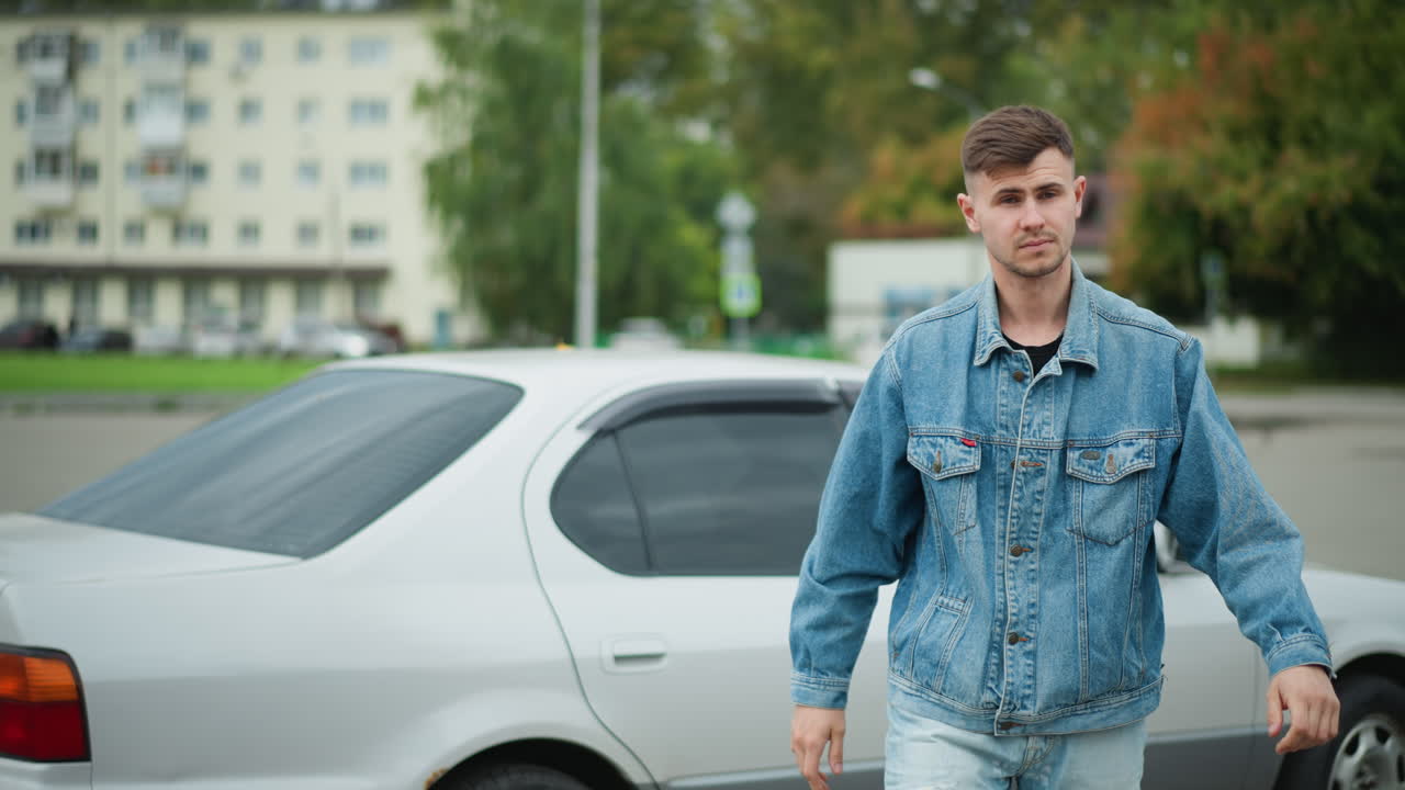 Uomo bianco alla guida di una berlina argentata esce da un parcheggio di periferia, giacca di jeans e jeans, apre la portiera, scende con passo sicuro, breve occhiata verso la strada, cielo nuvoloso, edificio sullo sfondo