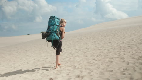 una excursionista rubia con una mochila pesada caminando descalza por una duna de arena en el desierto en un hermoso día de verano, råbjerg mile, dinamarca