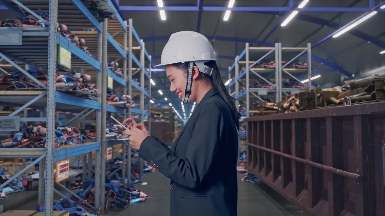 Вид сбоку: азиатская бизнесвумен использует планшет, идя по промышленному складу металлолома и вторсырья.