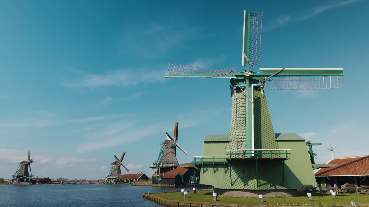 Ampia inquadratura panoramica degli iconici mulini a vento olandesi che costeggiano il fiume Zaan a Zaanse Schans, Paesi Bassi, in una soleggiata giornata di primavera sotto un cielo azzurro e limpido.