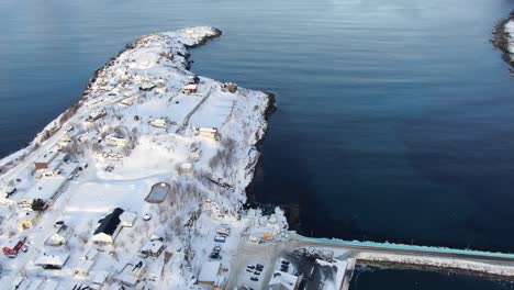 Drone-view-on-the-Tromso-mountains-in-winter-full-of-snow-showing-Husoy-a-small-town-on-an-island-surrounded-by-the-sea-and-its-small-port-with-flying-seagulls