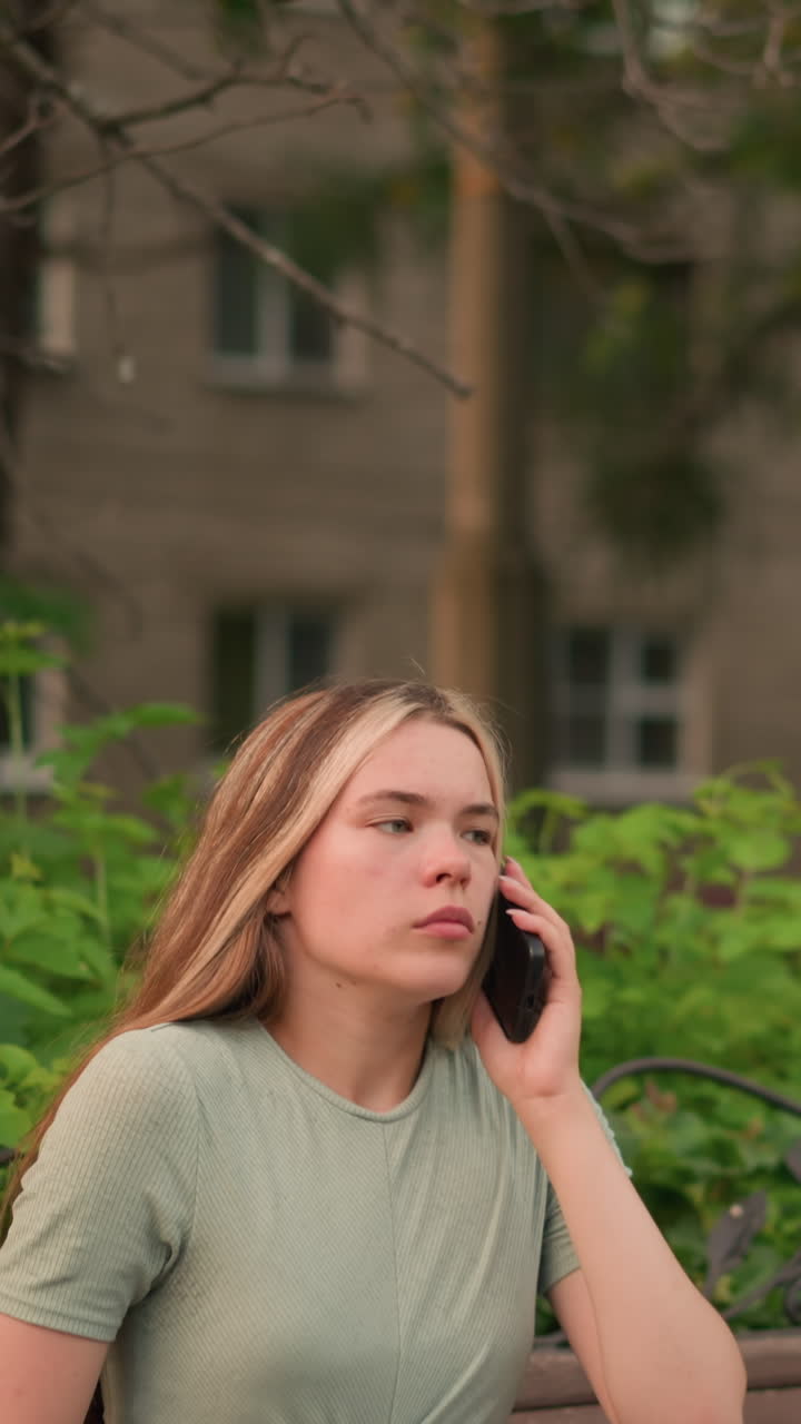 젊은 여성은 나무 벤치에 앉아 전화 통화를 하고, 머리카락을 조정하고 목에 손을 고, 울창한 초록색, 나무, 그리고 배경의 건물로 둘러싸여 있습니다.