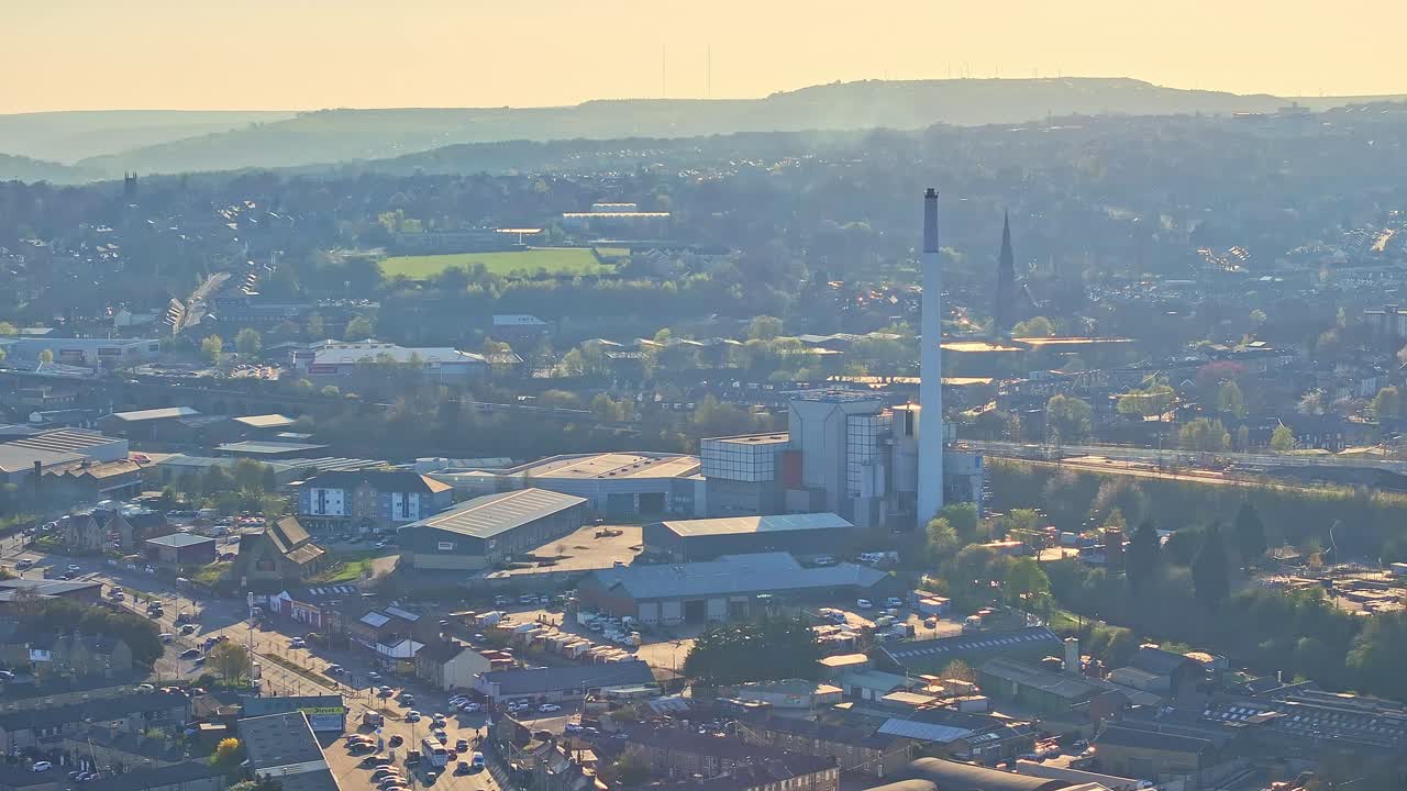 Drone captures sprawling industrial area near Huddersfield’s Springwood zone, including tall smokestacks, warehouses, and distant residential districts under soft evening light, aerial view, real time