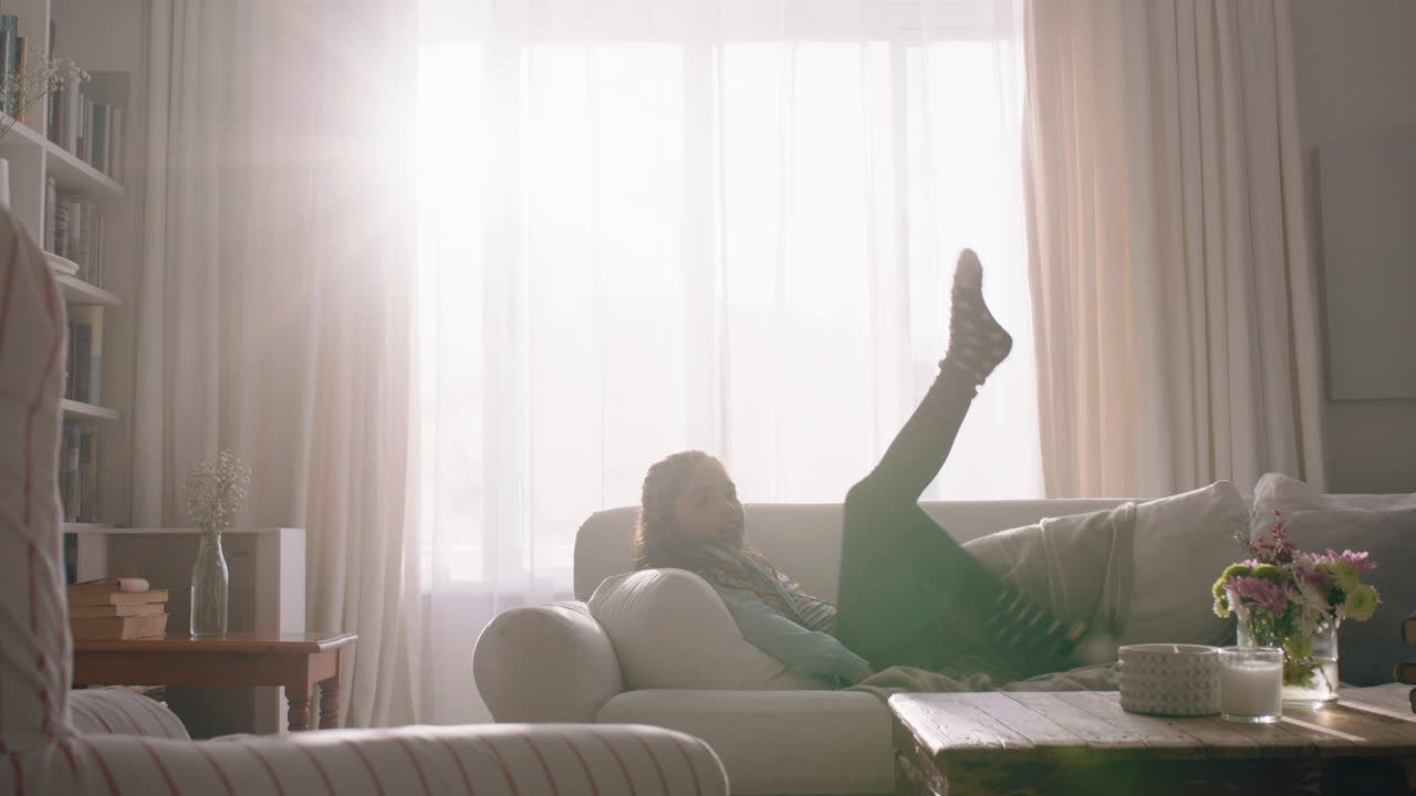 소파에 누워서 즐겁게 놀고 있는 행복한 어린 소녀 주말 아침을 즐기고 집에서 태양빛이 창문을 통해 빛나는 4k 영상
