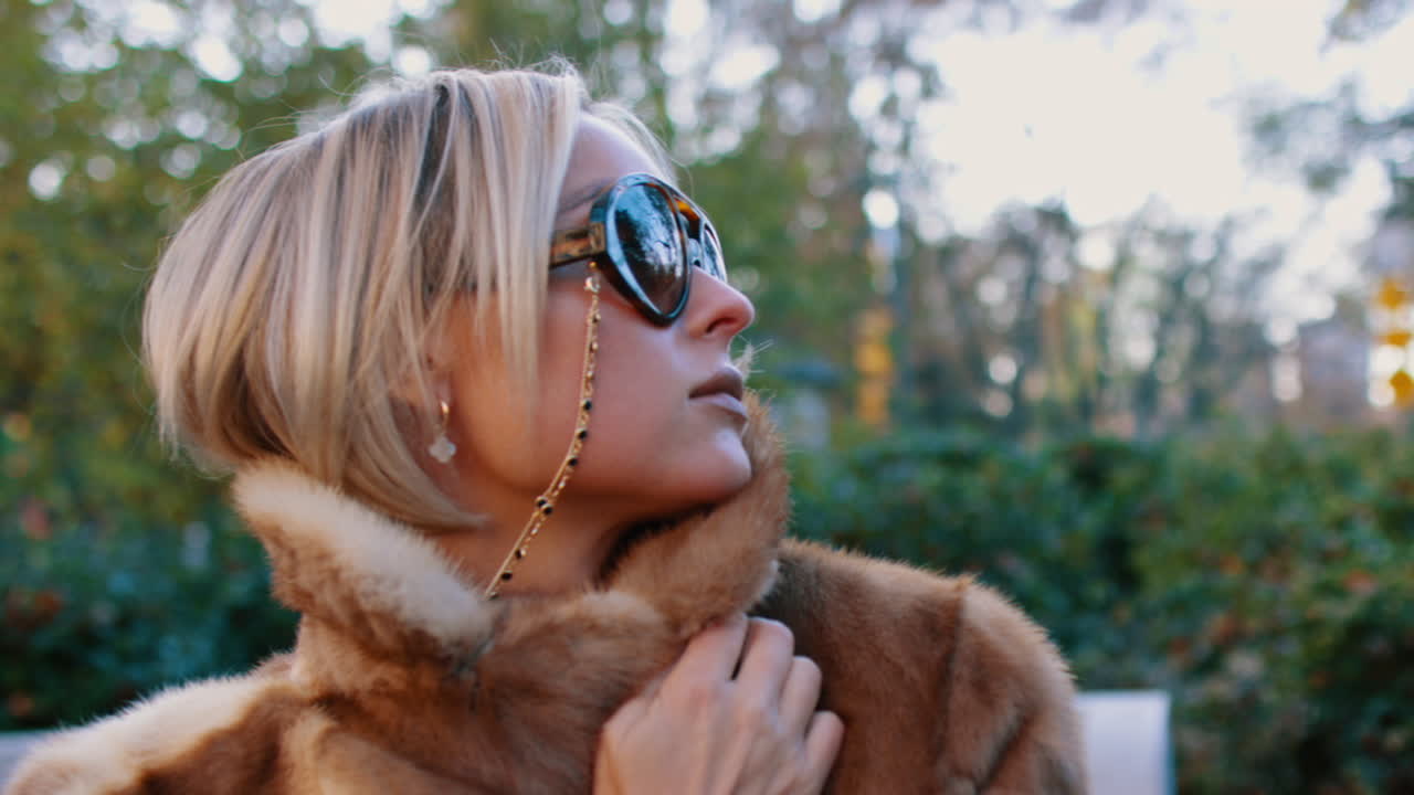Ritratto dal basso di donna in pelliccia che guarda in alto. Primo piano di donna bionda con occhiali da sole aviator, catena d'oro e colletto di pelliccia, sullo sfondo di alberi sfocati a New York.