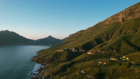 Hout-Bay-coastline-with-road-carved-into-Chapmans-Peak-at-sunset,-aerial