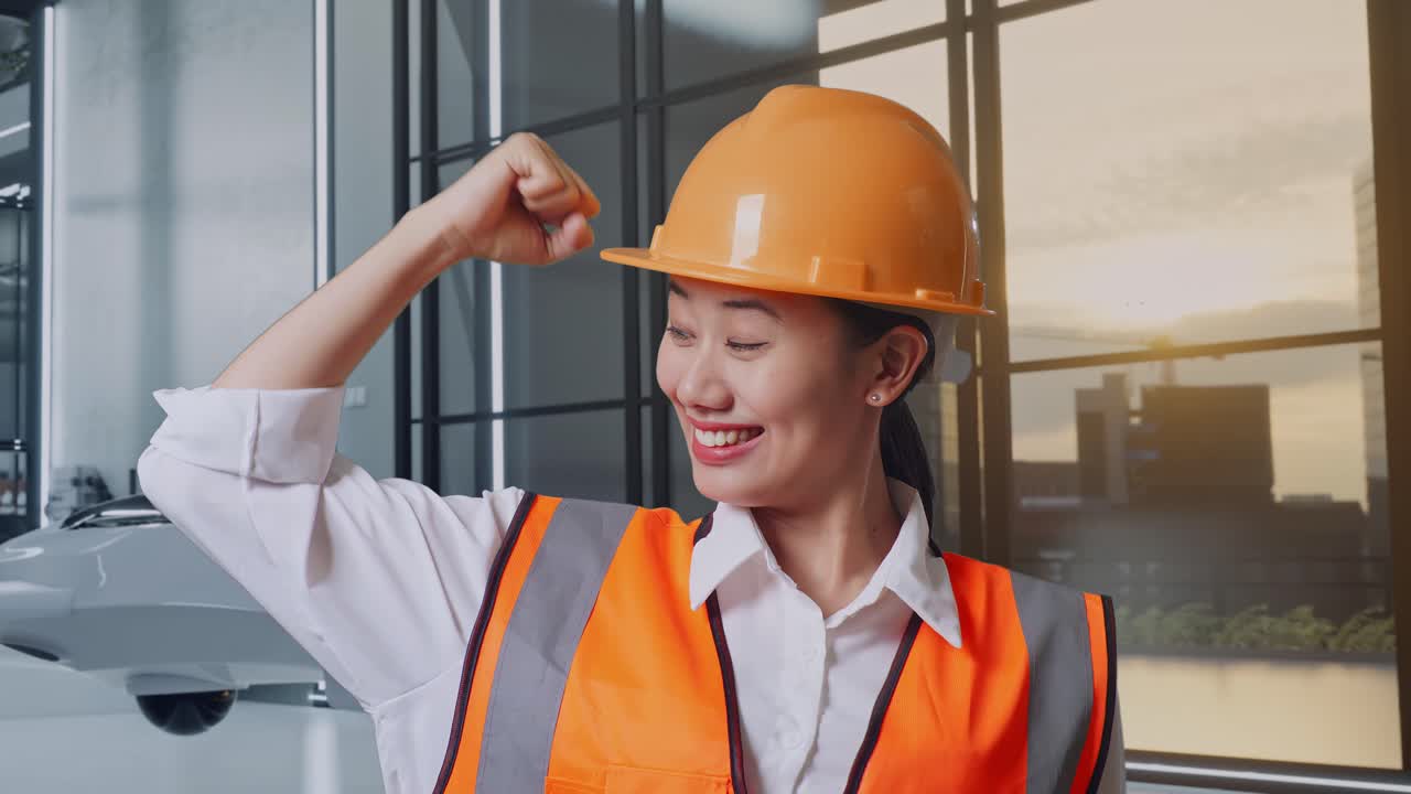 안전 헬을 착용한 아시아 여성 엔지니어가 이를 구부리고 드론 프로젝트에 서서 카메라를 향해 미소 짓고 있습니다.