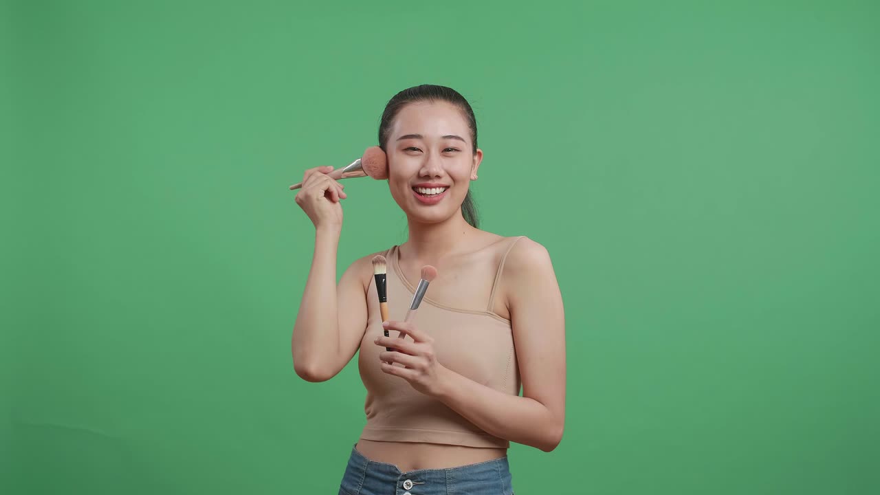 Beautiful Asian Woman Holding Makeup Brushes And Rouging Her Cheeks While Smiling To Camera On Green Screen Background In The Studio