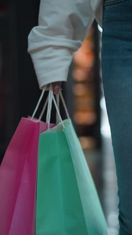 bolsa de compras rosa sostenida en la mano de una dama con uñas pulidas, complementada por una bolsa de color menta, puesta contra un vibrante fondo de un centro comercial con pantallas modernas y iluminación ambiental brillante