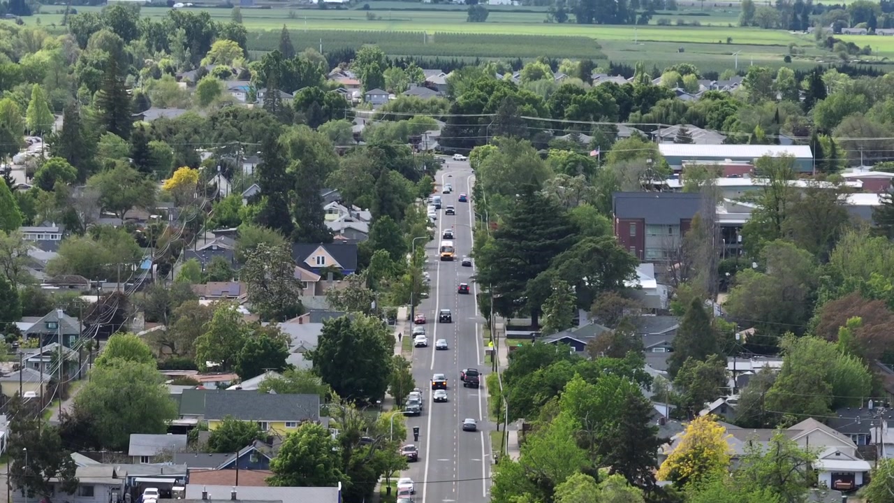 アメリカ合衆国オレゴン州メドフォードの住宅街を、並木道の大通りが交通量のある車、家々、春の木々を伴い、薄曇りの空の下を貫いている。空撮ドローン降下ショット。