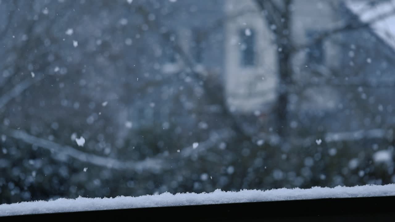 Gorgeous cinematic footage of white fluffy thick and dense snowflakes falling in slow motion behind a balcony during a cloudy day with a large tree and building in the background blurred out.
